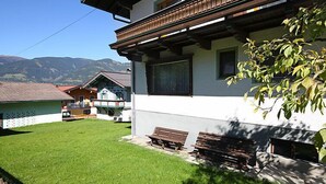 Un edificio de dos pisos con balcón, un patio césped y montañas al fondo.
