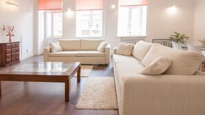 Ein geräumiges Wohnzimmer mit einem beige farbenen Sofa, einem Holztisch und einem großen Fenster mit orangefarbenen Jalousien.