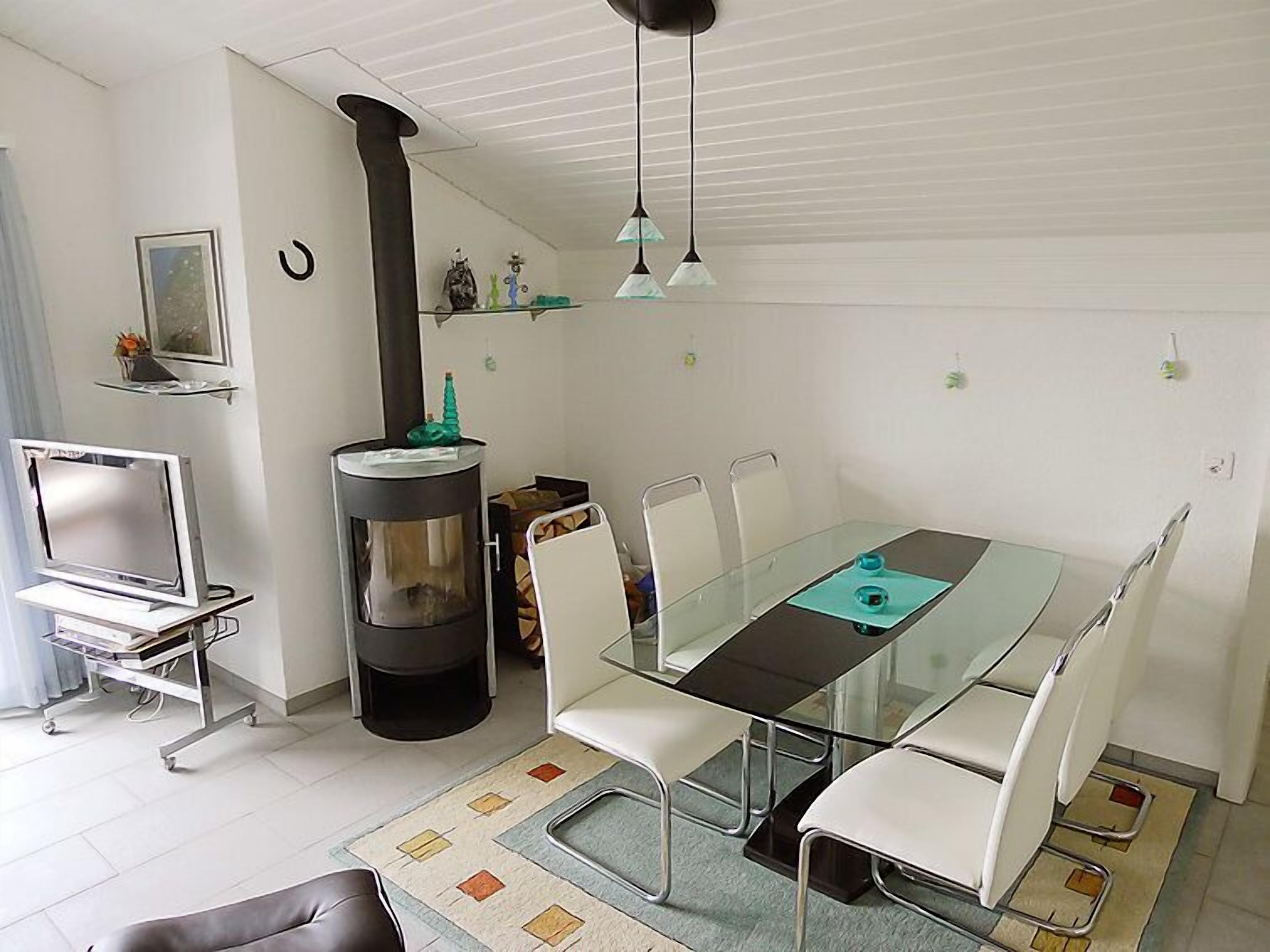 Ein modernes Wohnzimmer mit einem Glastisch, weißen Stühlen, einem schwarzen Kamin und einem Fernseher auf einem Schrank.