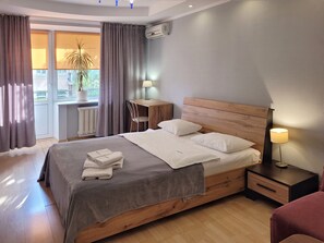 Ein ordentlich bezogenes Bett mit Kissen und Decke, ein Nachttisch mit Lampe, ein Fenster mit Vorhängen und eine Heizung.