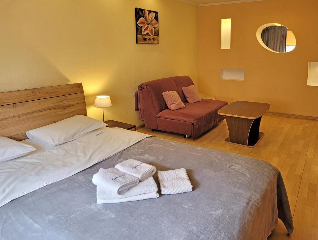 Une chambre d’hôtel avec un lit doté d’une tête de lit en bois, une couvre-lit grise et un canapé rouge avec deux coussins. Une petite table et une lampe se trouvent sur le côté droit.