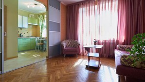 Un salon doté d’un parquet en chevrons, d’un canapé, d’un fauteuil, d’une petite table et d’une vue sur la cuisine, accessible par une porte-fenêtre coulissante.