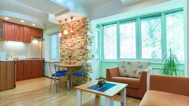 A modern kitchen with wooden cabinets, a dining area with a table and chairs, a living room with a sofa and a coffee table, and large windows with a view of trees. - Home-Hotel Metropolita Sheptitskogo 1A (Kyiv)