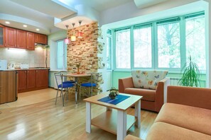 Una cocina moderna con armarios de madera, un comedor con mesa y sillas, una sala de estar con sofá y una mesa de centro, y ventanas amplias con vistas a árboles.