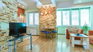 A modern living room with a stone wall, a flat-screen TV on a glass table, a round dining table with blue chairs, a sofa, and a potted plant. - Home-Hotel Metropolita Sheptitskogo 1A (Kyiv)