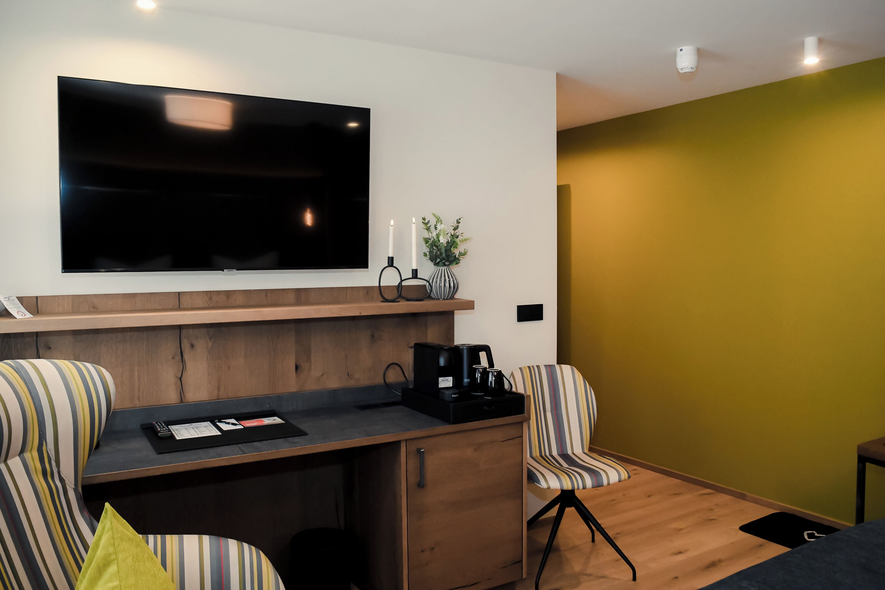 A modern room with a flat-screen TV mounted on the wall, a wooden desk with a coffee maker, striped armchairs, and a small plant.
