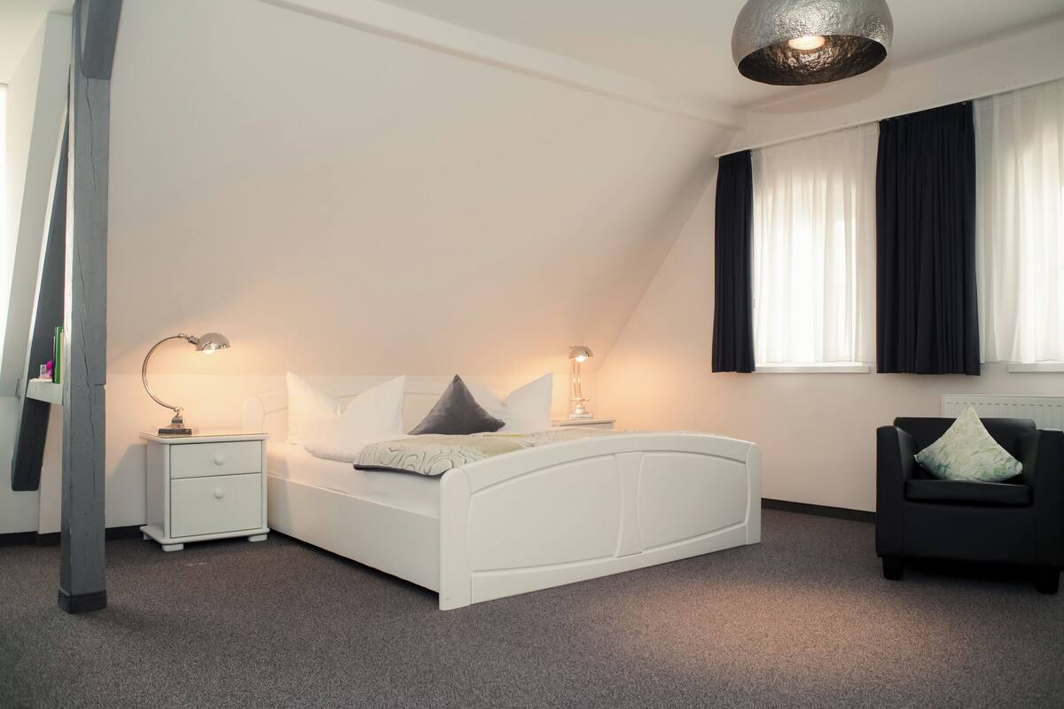 A bedroom with a white bed, two bedside tables with lamps, a black armchair, and a large window with black curtains.