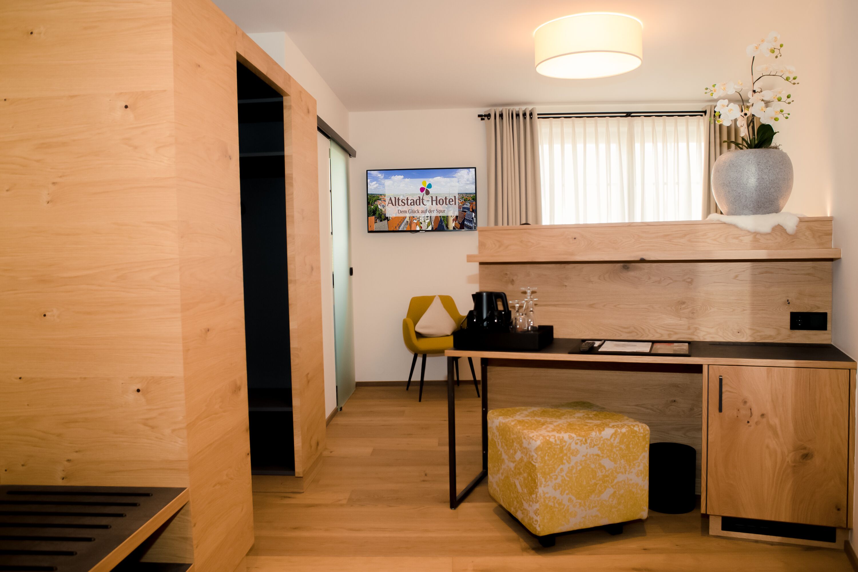 Una habitación compacta con un escritorio de madera, una silla amarilla, un pequeño reposapiés, un televisor montado en la pared y una ventana con cortinas.