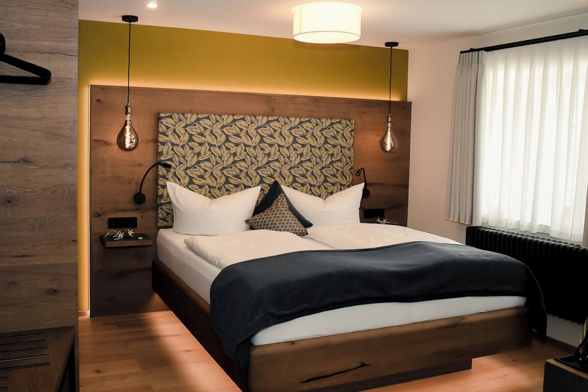 A modern bedroom with a wooden headboard, a bed with white bedding and a dark throw, bedside tables with lamps, and a window with white curtains.