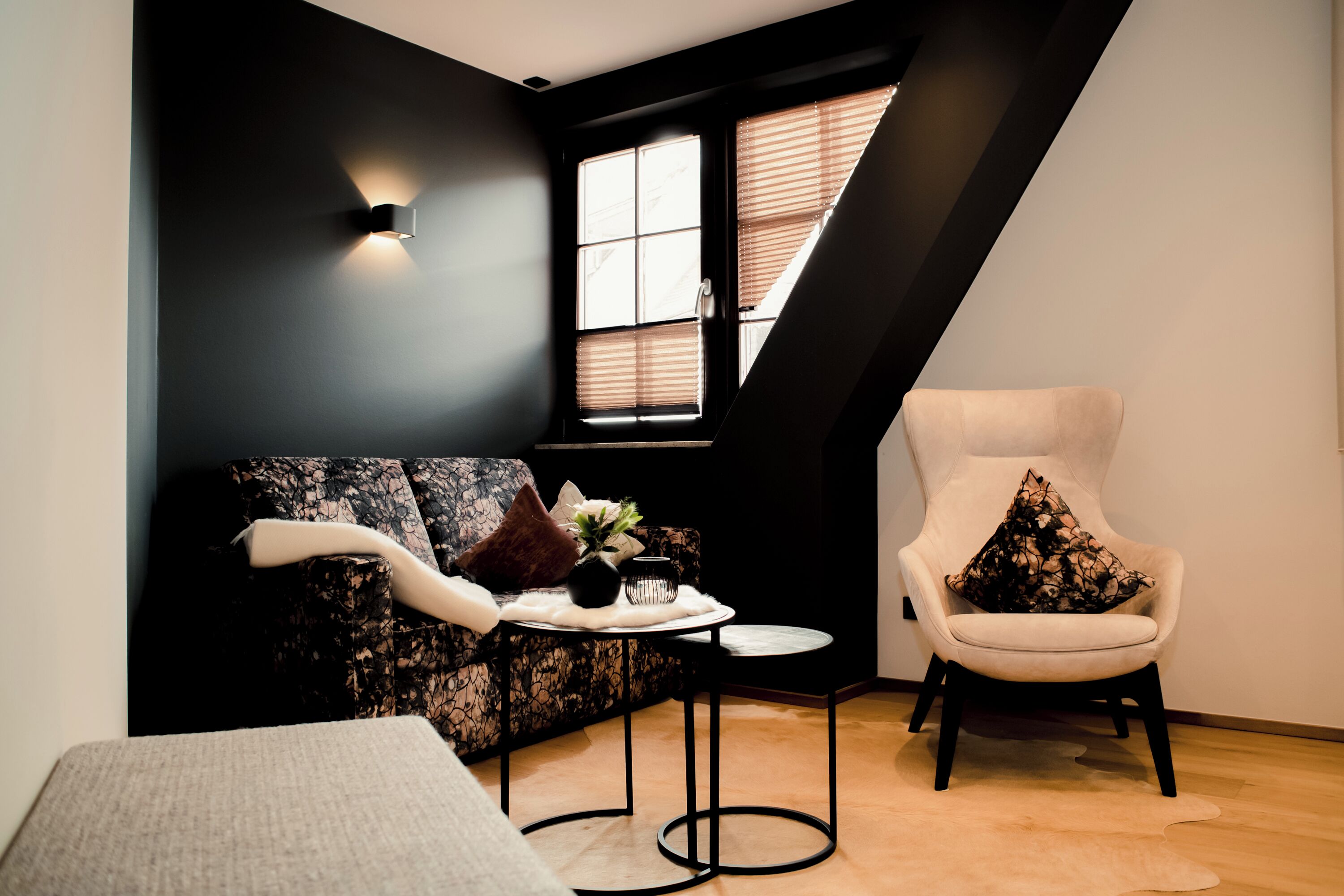 A modern loft bedroom with a dark accent wall, a window with blinds, a sofa with decorative pillows, a chair, and a small round table with a vase.