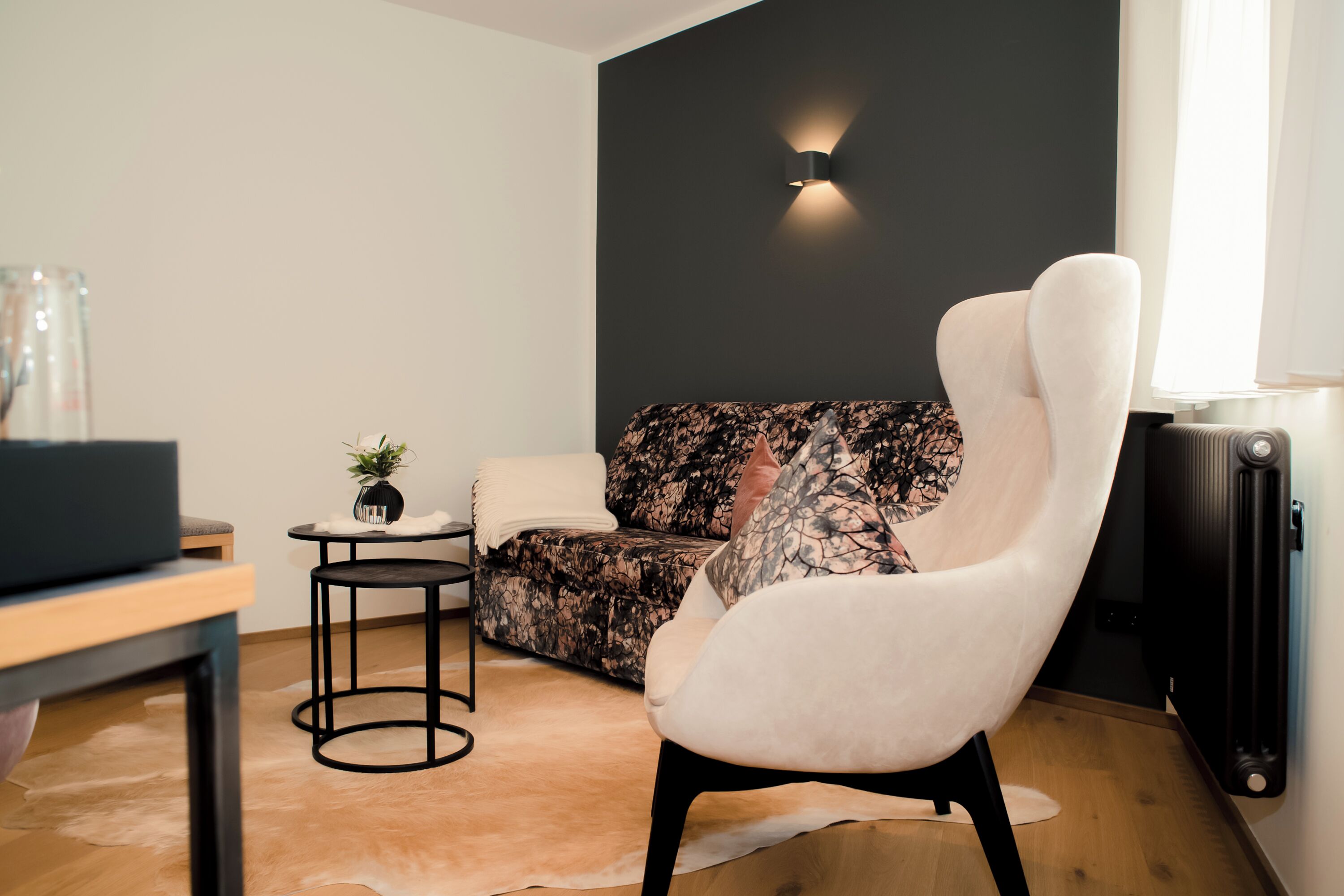 A modern living room with a sofa, a white armchair, a small side table with a plant, and a black wall-mounted light.