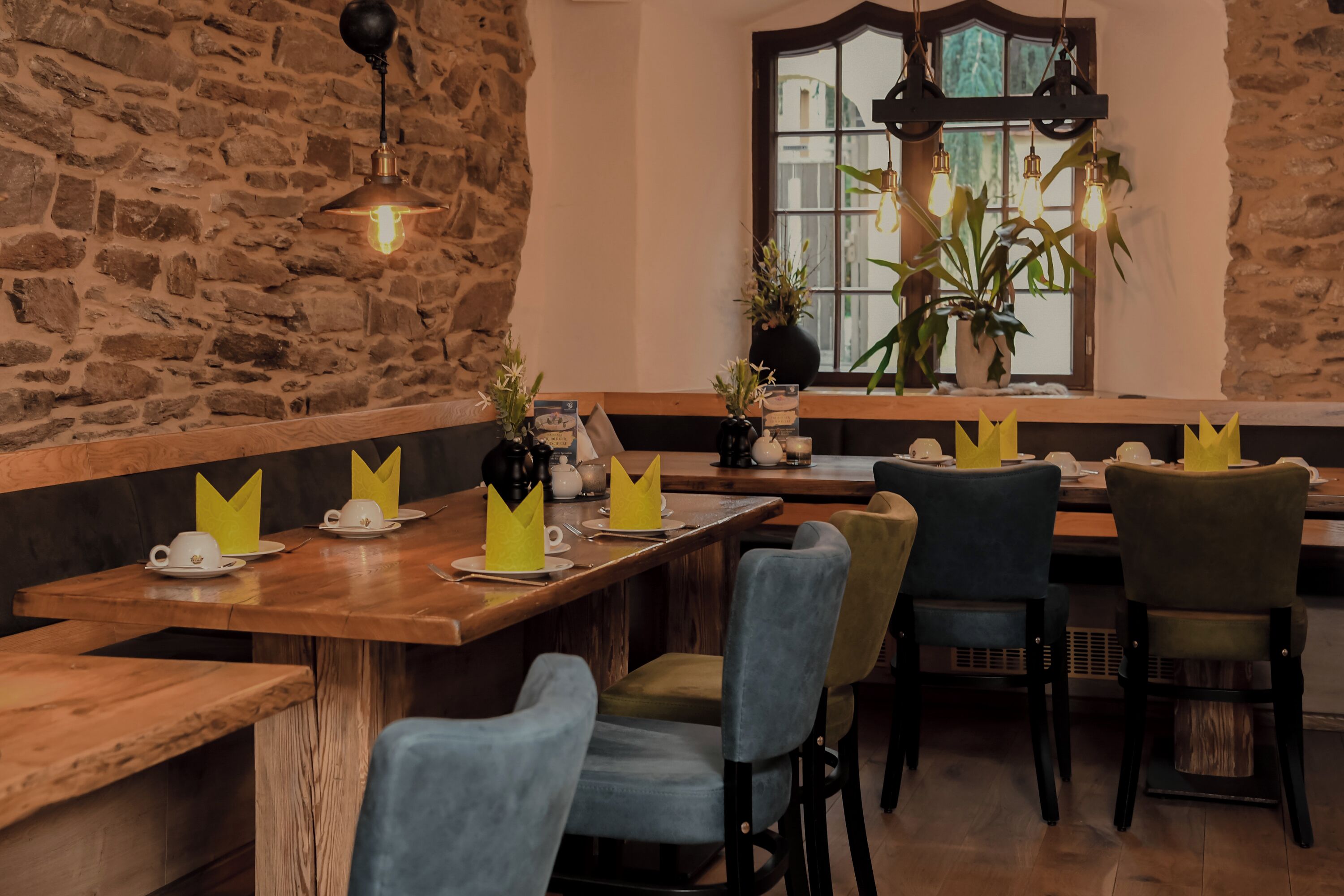 Um restaurante com mesas e cadeiras de madeira, parede de pedra e uma janela com plantas.