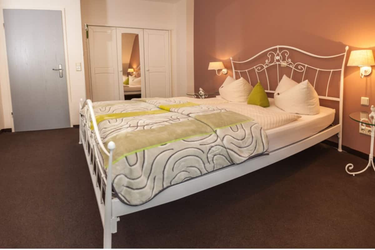 A neatly made bed with a white metal frame, a patterned bedspread, and several pillows. There is a bedside table with a lamp and a small decorative item. A door and a mirror are visible in the background.