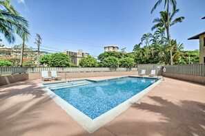 Une piscine rectangulaire dont les bords sont recouverts de carreaux, entourée d’une terrasse en béton. On y trouve deux chaises longues et un petit escalier pour entrer dans la piscine. L’ensemble est délimité par un mur bas, avec un palmier et d’autres plantes en arrière-plan.