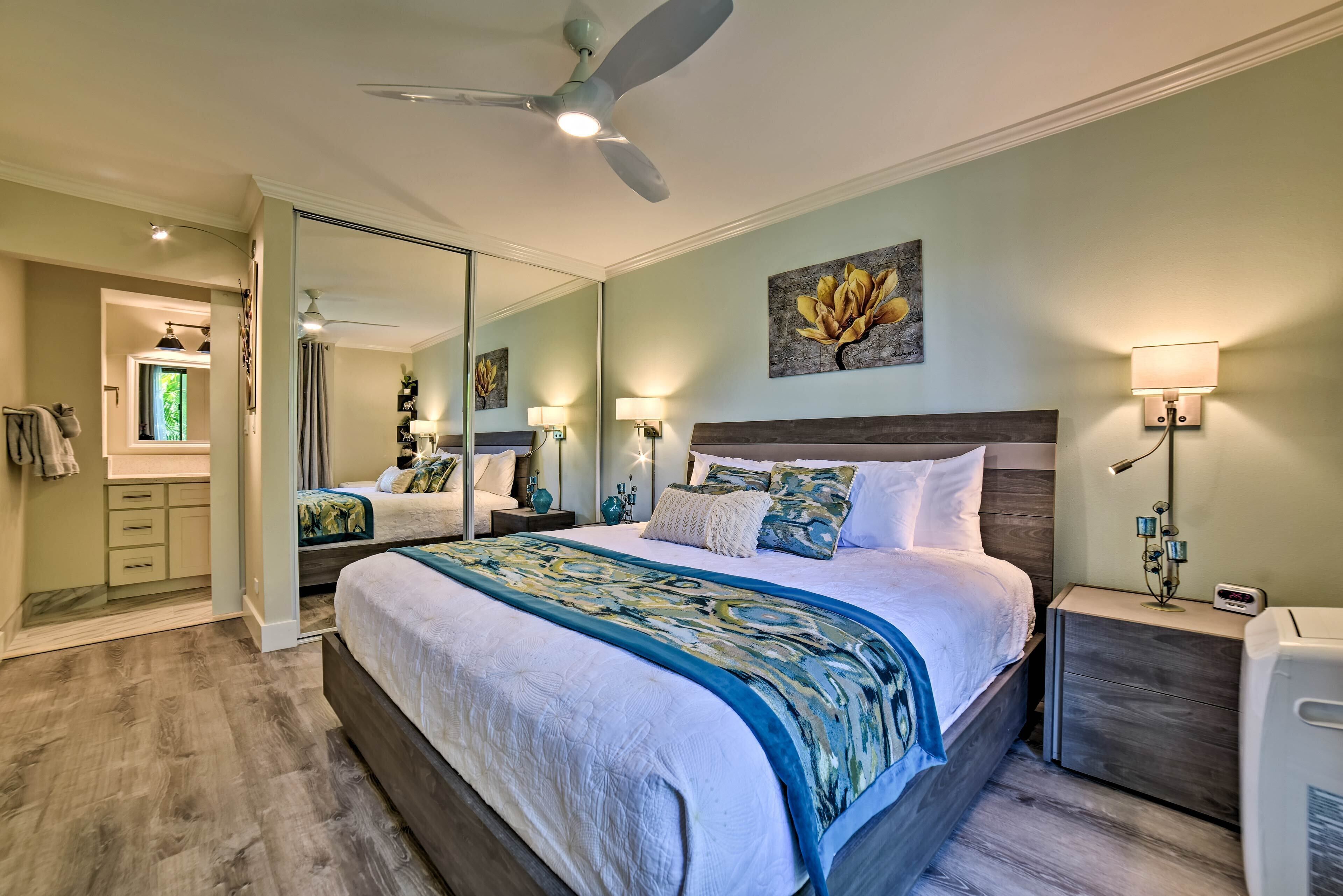 A bedroom with a large bed, a ceiling fan, a nightstand with a lamp, a mirror, and a painting on the wall.
