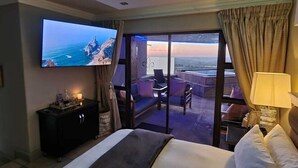 Een hotelkamer met een groot bed, een televisie aan de muur, een balkon met uitzicht op zee en een bureau met een lamp.