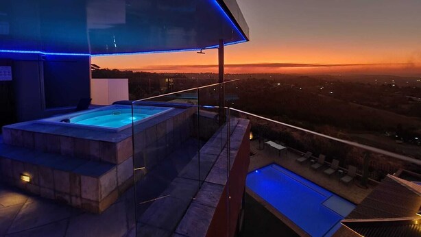 Een dakzwembad met een hottub en een kleiner zwembad, omgeven door glazen balustrades en blauwe LED-verlichting.