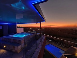 Un espace piscine sur le toit, comprenant un jacuzzi et une piscine plus petite, le tout entouré de rampes de verre et illuminé par des lumières LED bleues.