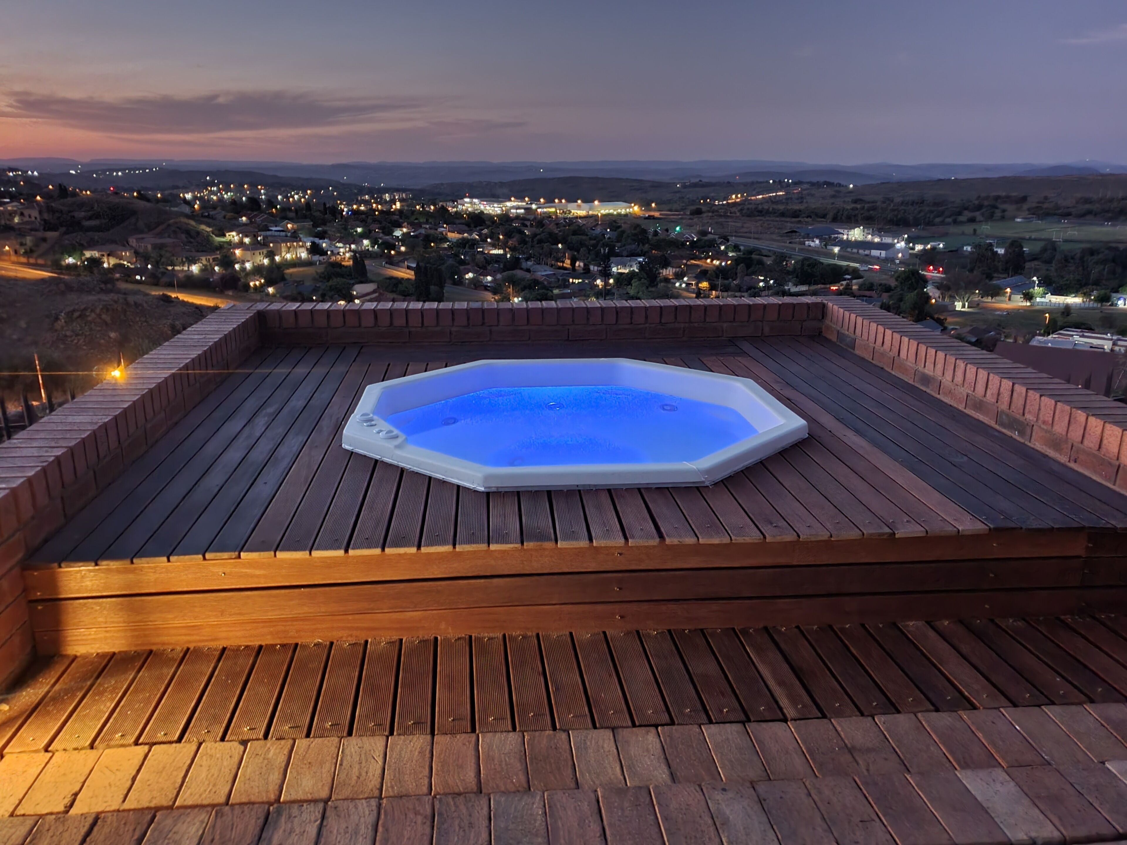 A hot tub on a wooden deck with a view of a town at dusk.