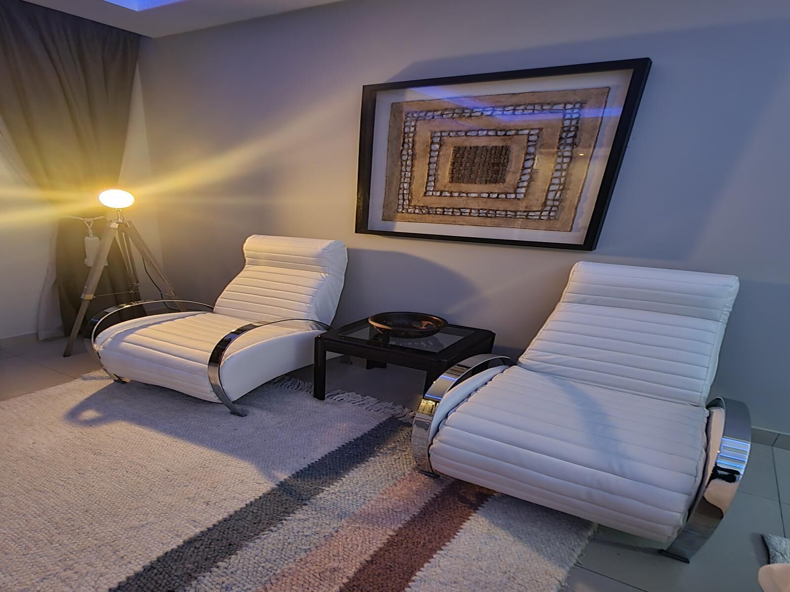A modern living room with two white leather armchairs, a small black side table, a framed abstract artwork, and a floor lamp.