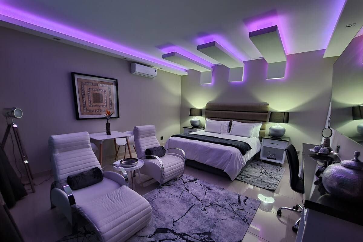 Een moderne slaapkamer met een bed, twee stoelen, een klein tafeltje en een nachtkastje met een lamp.