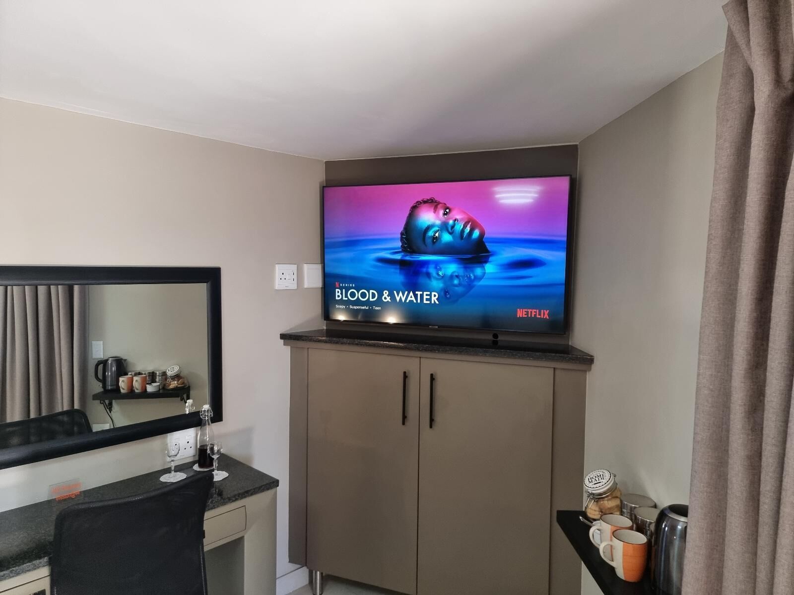 A modern hotel room with a flat-screen TV mounted on a cabinet, a desk with a chair, and a mirror above the desk.