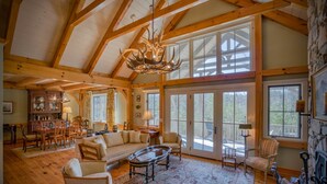 A spacious living room with a high ceiling, wooden beams, a chandelier, a fireplace, and large windows. - Elegant Mountain Dream Escape: 3BR with Fireplace & Views (Mill Spring)