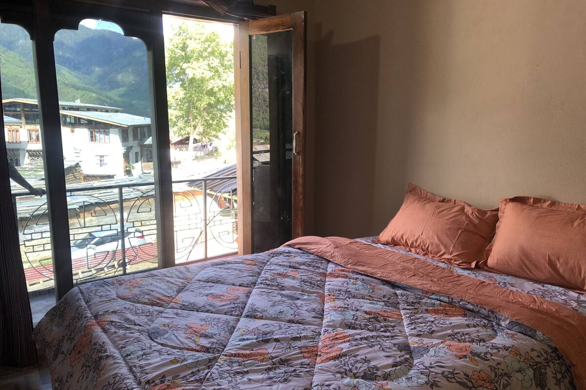 A bedroom with a bed, a window, and a view of buildings and trees.