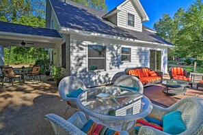 Une terrasse couverte avec des meubles en osier et une table en verre, un porche couvert avec des chaises, et une maison avec un toit foncé et un revêtement blanc.