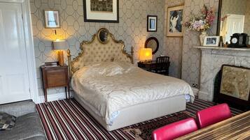 A bedroom with a large bed, a nightstand with a lamp, a dresser, a mirror, and a fireplace.