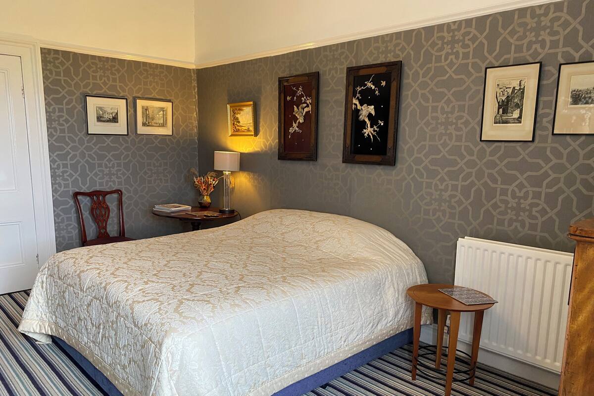 Un dormitorio ordenado con una cama grande, una mesita de noche con lámpara, una silla y cuadros enmarcados en la pared.