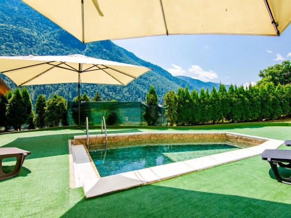 A swimming pool with a mountain backdrop, surrounded by green artificial turf and shaded by large umbrellas.