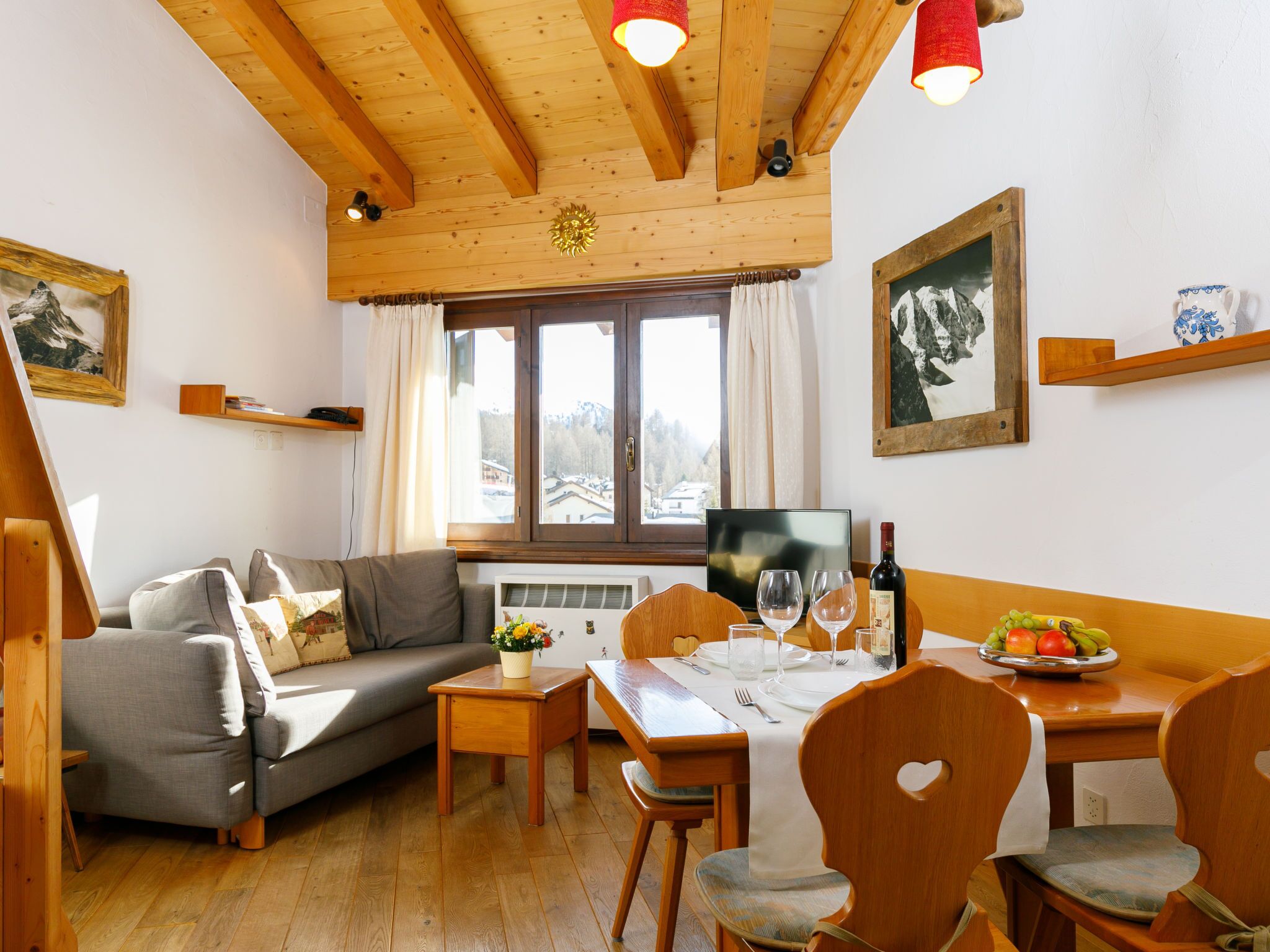 Un salon chaleureux avec un plafond en bois, un canapé gris, une table à manger dressée pour deux personnes et une grande fenêtre offrant une vue sur une montagne.