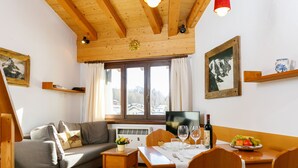 Ein gemütliches Wohnzimmer mit Holzbalkendecke, einem grauen Sofa, einem Esstisch für zwei Personen und einem großen Fenster mit Bergblick.
