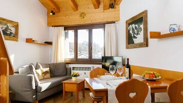 Ein gemütliches Wohnzimmer mit Holzbalkendecke, einem grauen Sofa, einem Esstisch für zwei Personen und einem großen Fenster mit Bergblick.