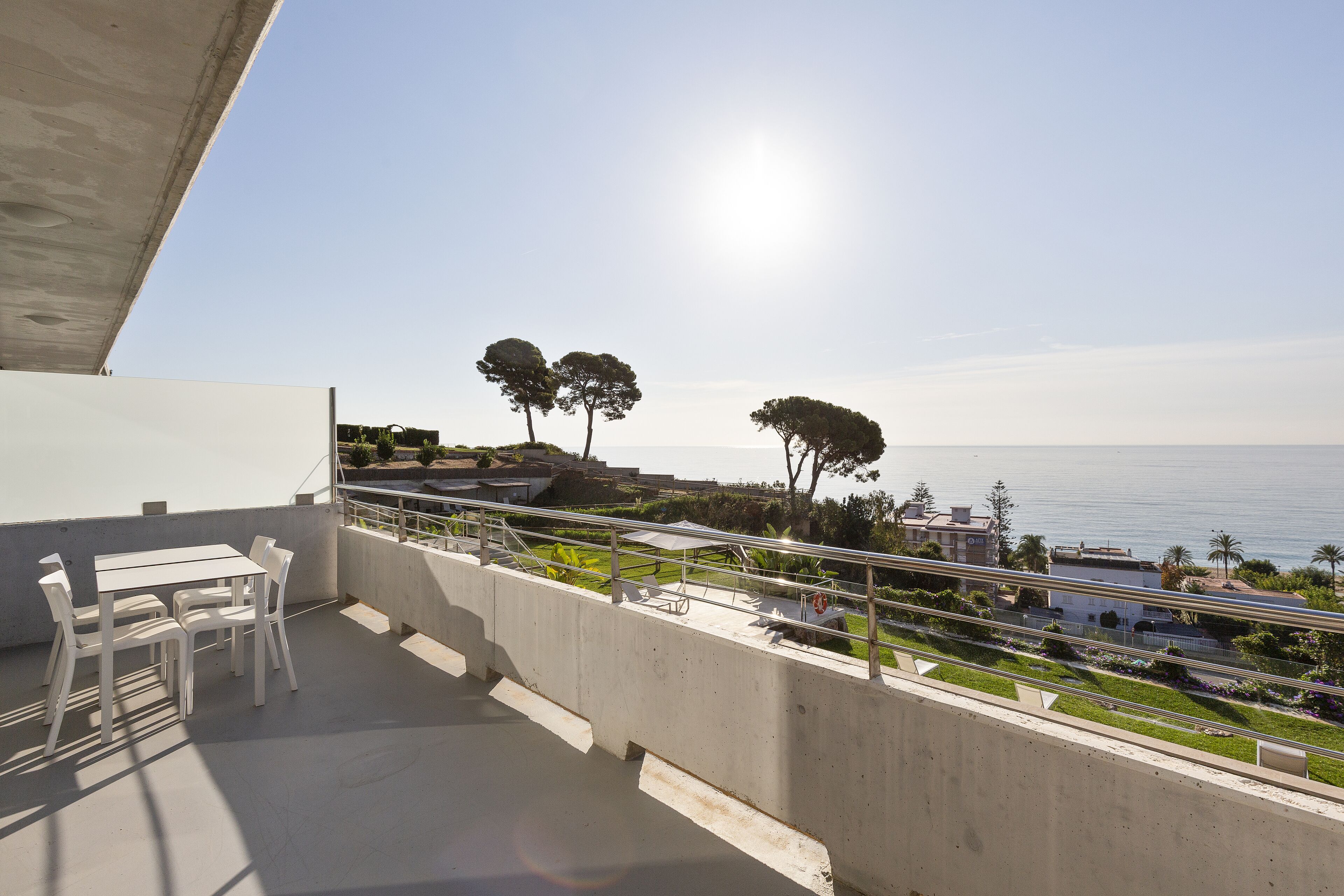 A balcony with a table and chairs, overlooking a coastal area with trees and a clear sky.