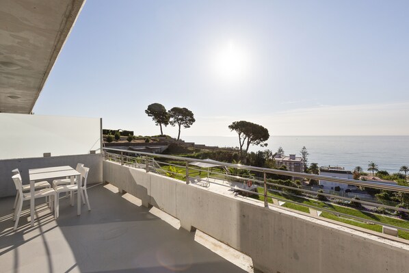 A balcony with a table and chairs, overlooking a coastal area with trees and a clear sky. - Santa Romana (Caldes d'Estrac)