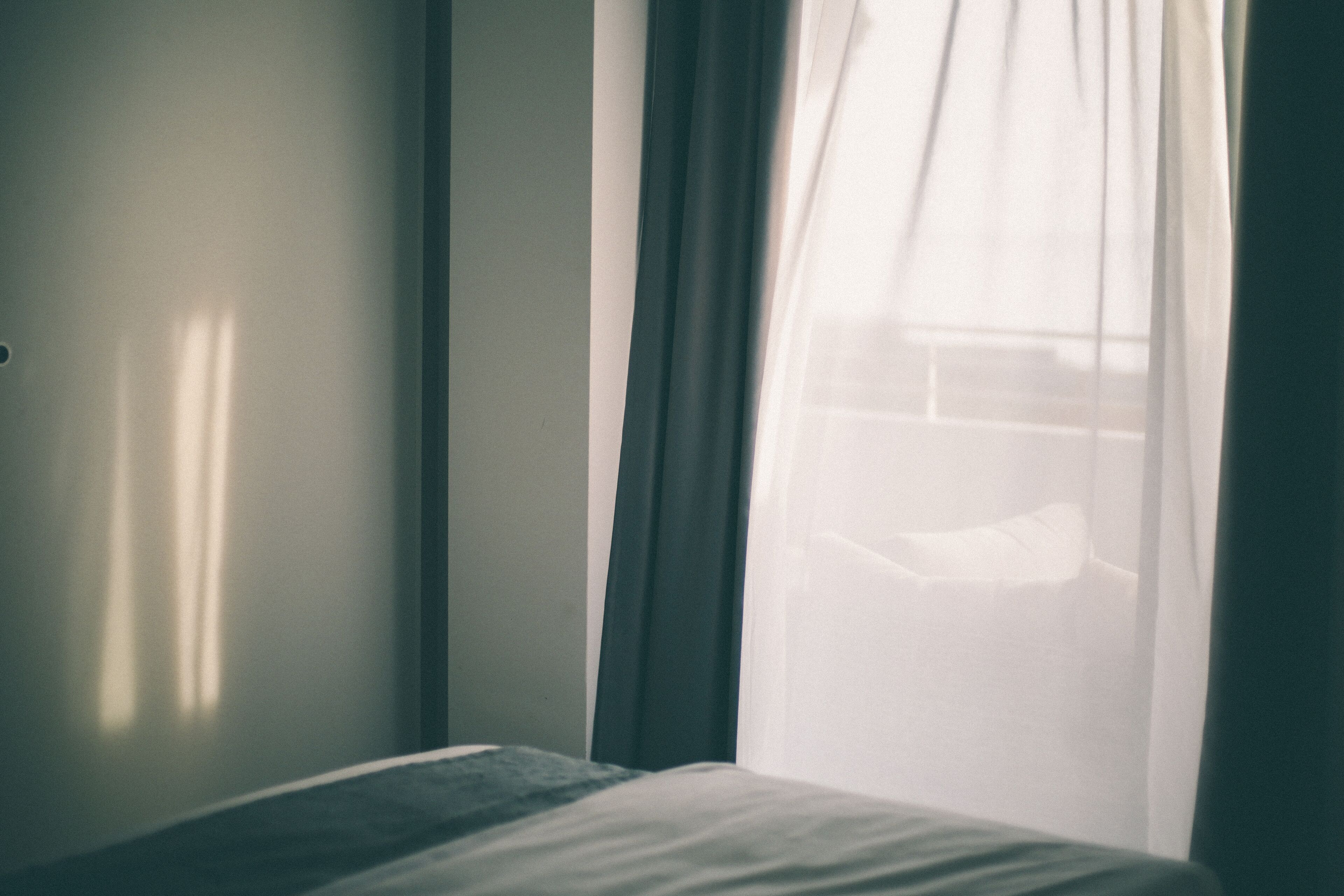 A room with a bed, a window with sheer curtains, and a wall with a light switch.
