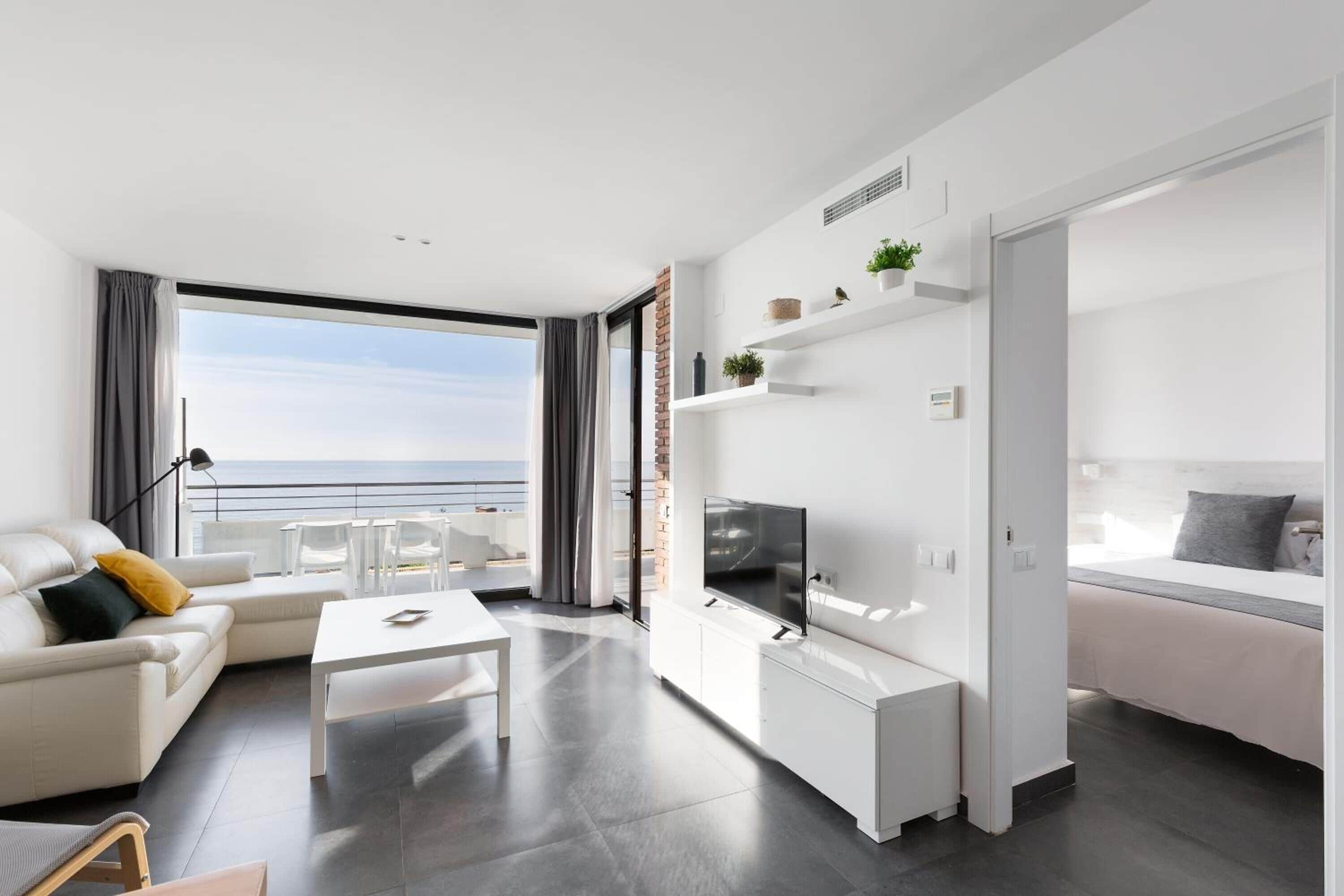 A modern living room with a sofa, coffee table, TV, and a balcony with a view of the sea.