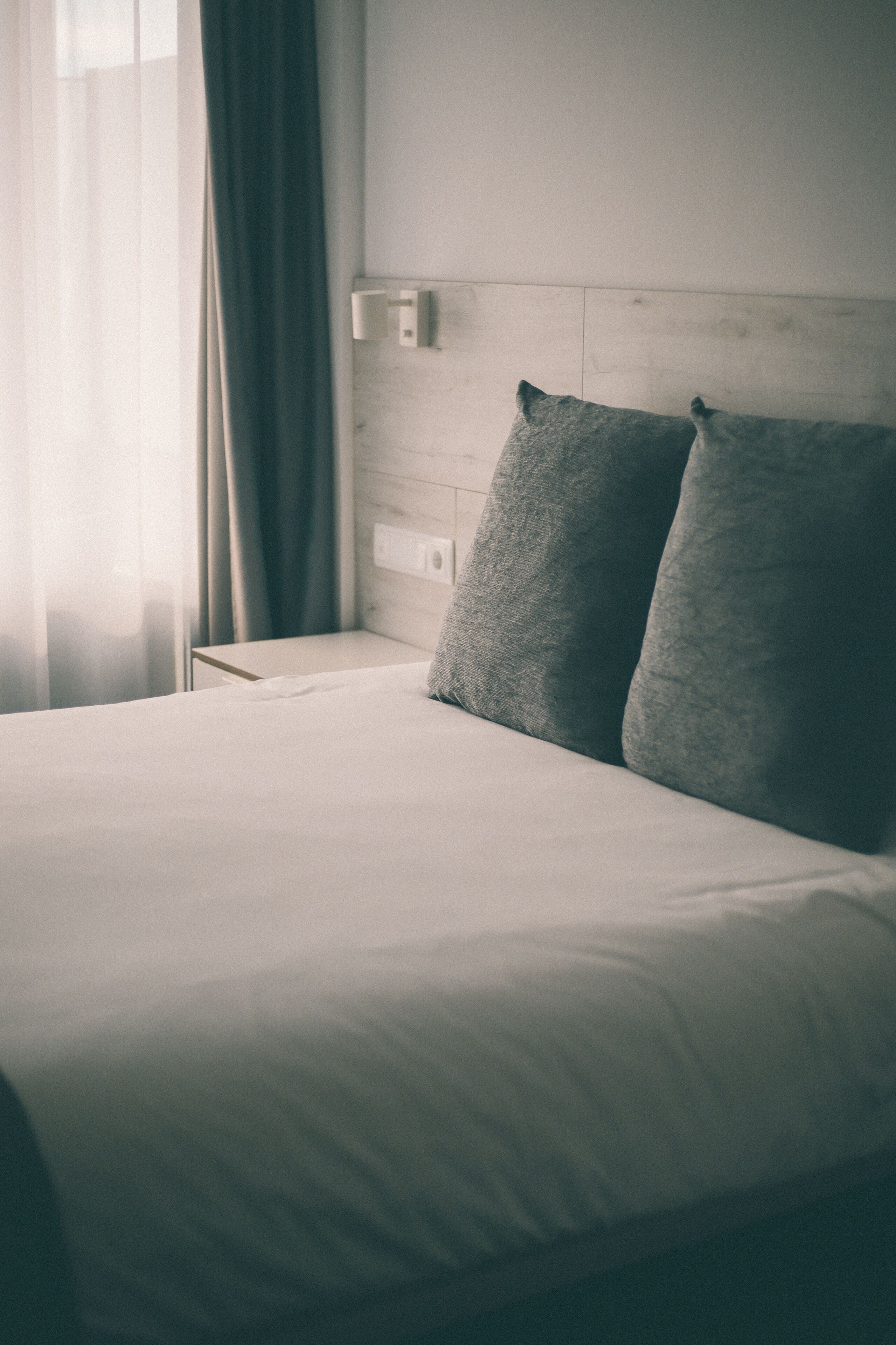 Una cama bien hecha con sábana blanca y dos almohadas, una mesita de noche con lámpara, y una ventana con cortinas.