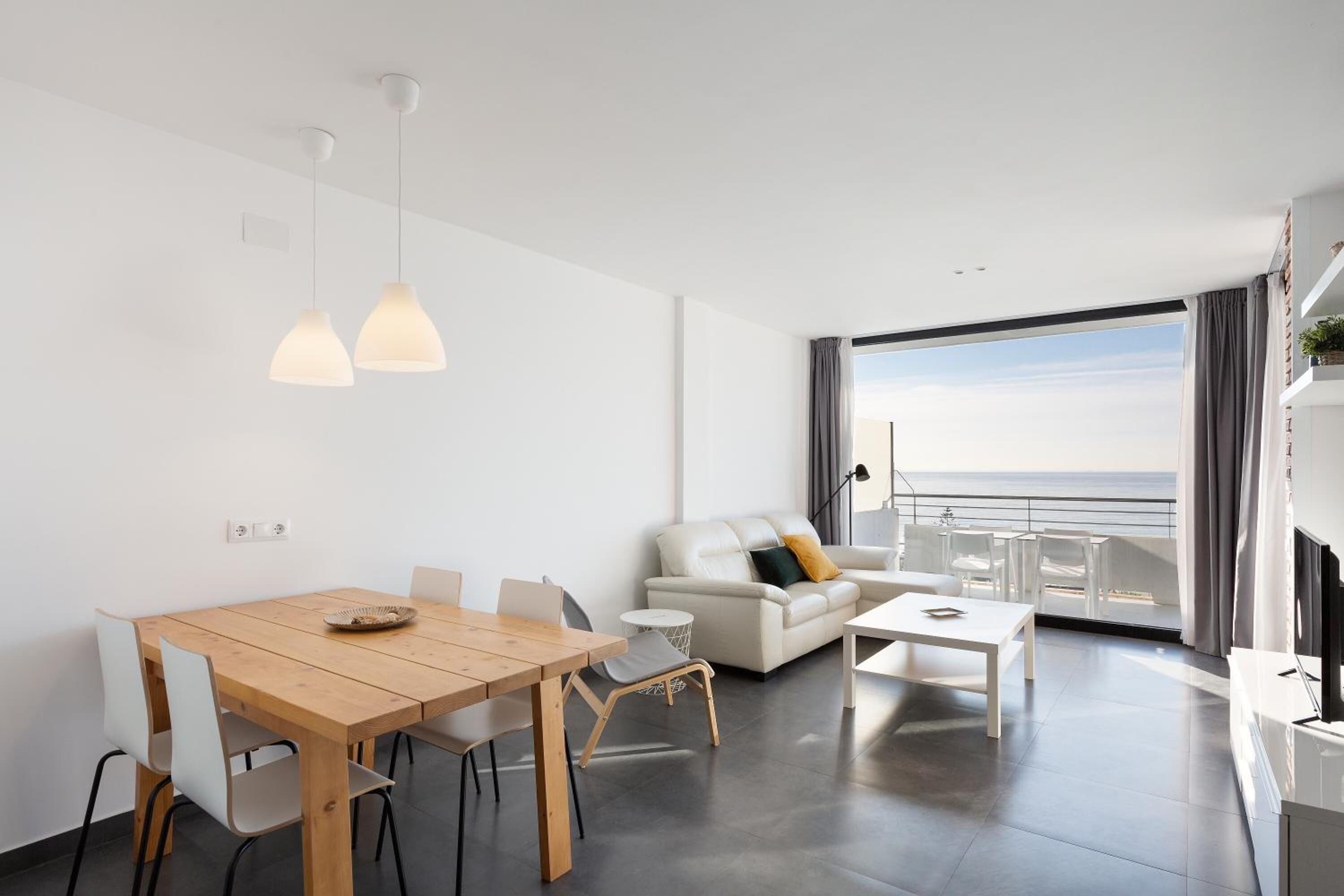 Una sala de estar moderna con una mesa de comedor de madera, sofás blancos y un balcón con vistas al mar.