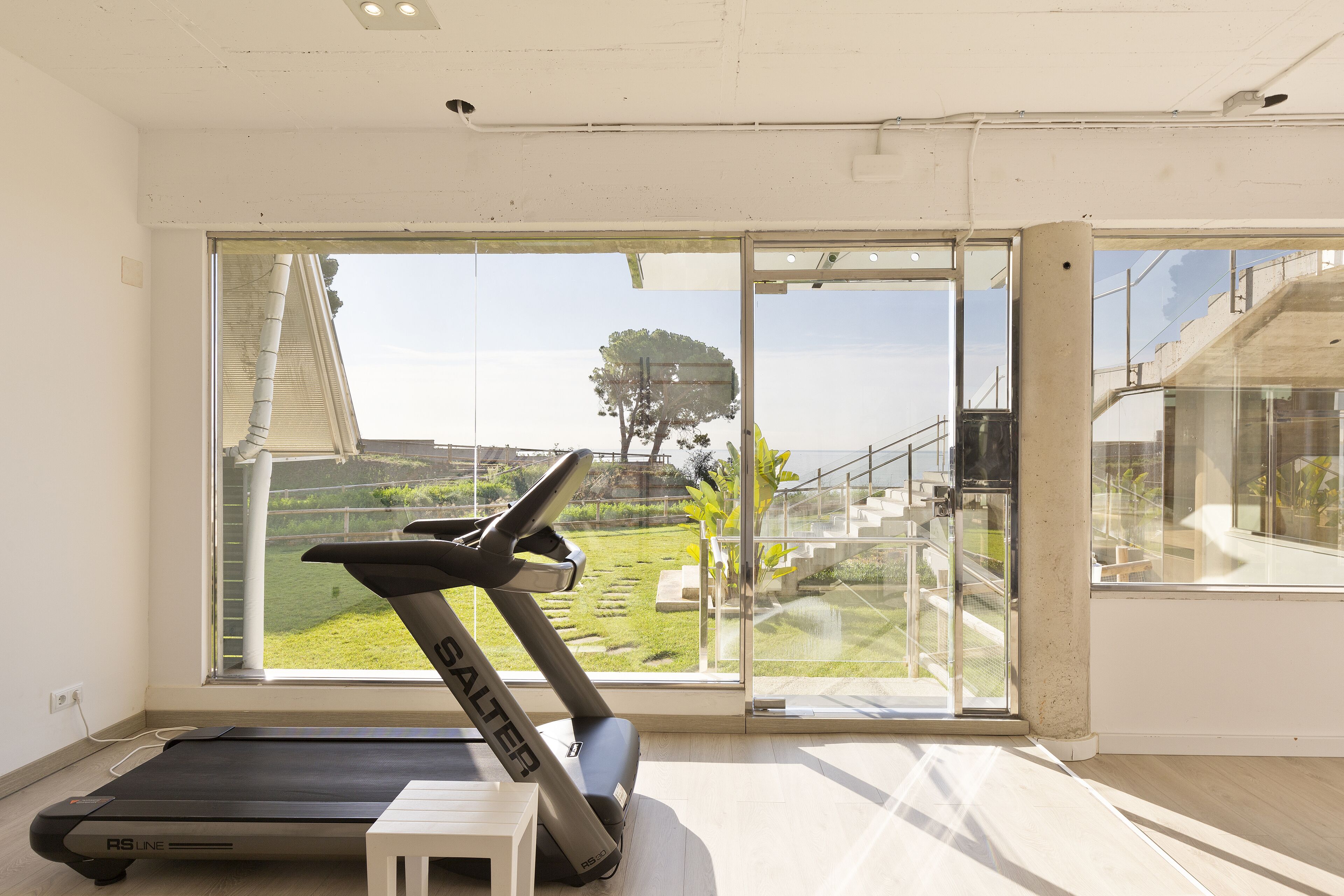 A home gym with a treadmill and a small white stool, large windows with a view of a garden and a tree.