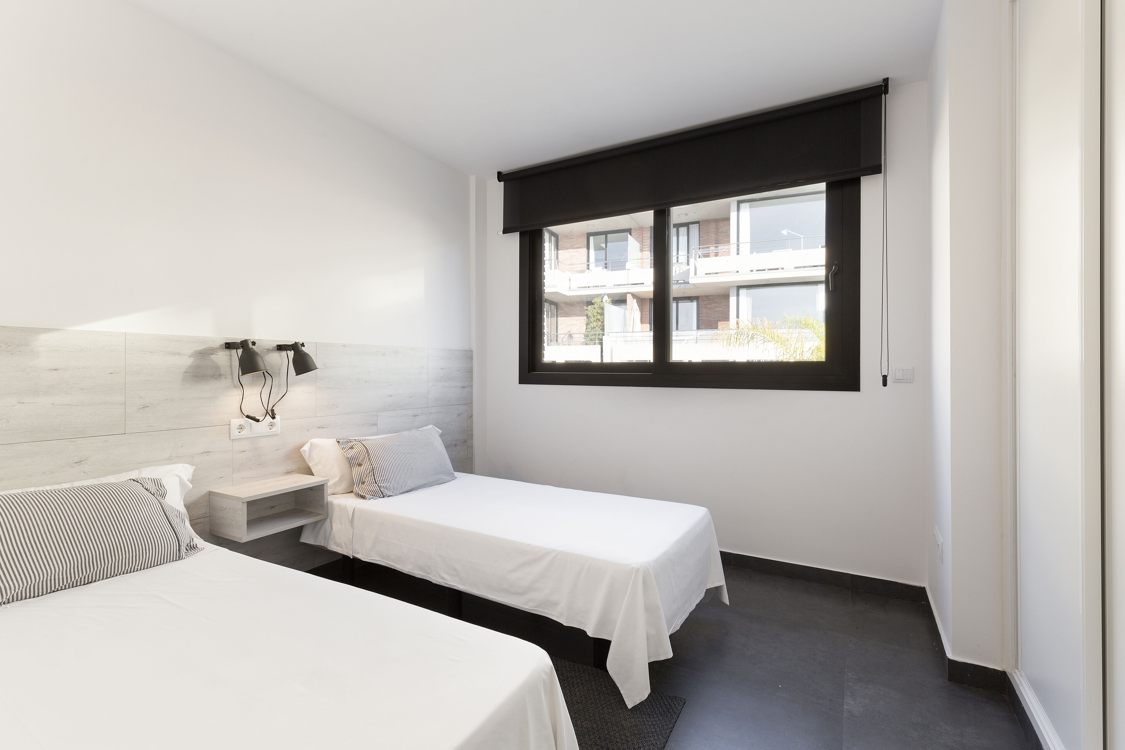 Una habitación de hotel moderna con dos camas individuales, un pequeño rincón de descanso, una ventana con vista al edificio y una lámpara fijada a la pared.