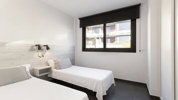 Una habitación de hotel moderna con dos camas individuales, un pequeño rincón de descanso, una ventana con vista al edificio y una lámpara fijada a la pared.