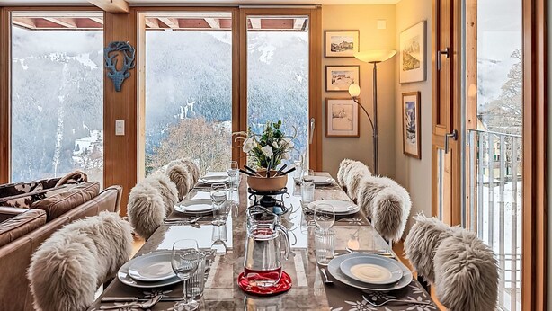 A dining room with a long table set for six, large windows offering a mountain view, wooden beams on the ceiling, and framed artwork on the walls. - Roossi Apt. 1 by Interhome (Wengen)