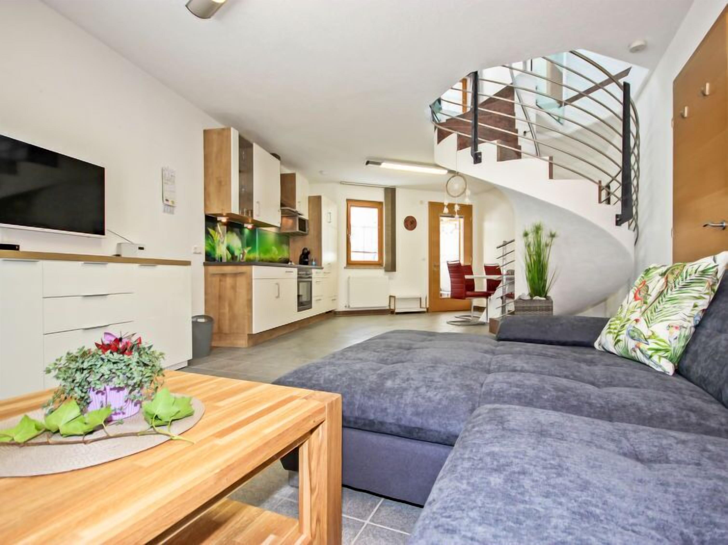 Un salon moderne avec un escalier à la forme arrondie, un téléviseur à écran plat, une table à manger en bois agrémentée d’une plante et un canapé gris.