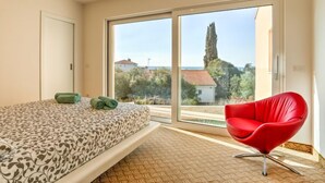 A bedroom with a bed, a red chair, and a large sliding glass door leading to a balcony with a view of houses and trees. - Apartment Karmen by Interhome (Umag)