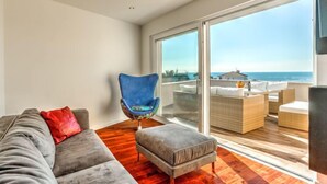 A modern living room with a grey sofa, a blue chair, and an ottoman. It features a large sliding glass door leading to an outdoor seating area with a view of the ocean. - Apartment Karmen by Interhome (Umag)