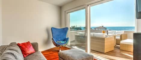 A modern living room with a grey sofa, a blue chair, and an ottoman. It features a large sliding glass door leading to an outdoor seating area with a view of the ocean.