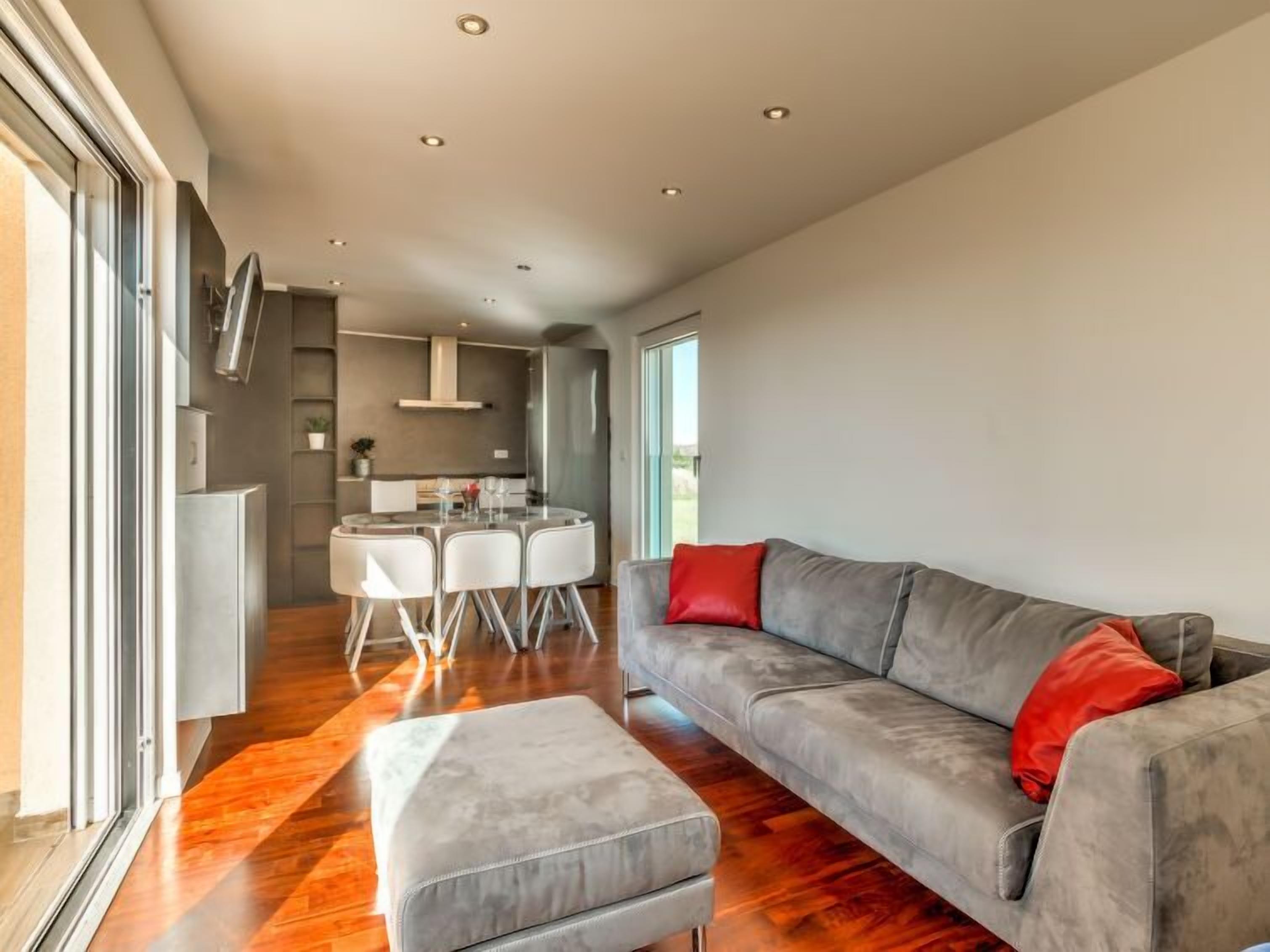 A modern living room with a grey sofa, a glass dining table, and a grey ottoman.